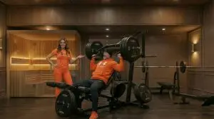 Perfect Home Gym Blog 77 Mann trainiert an Kraftmaschine, Frau zeigt auf ihn, Sauna mit Glasfront im Hintergrund.