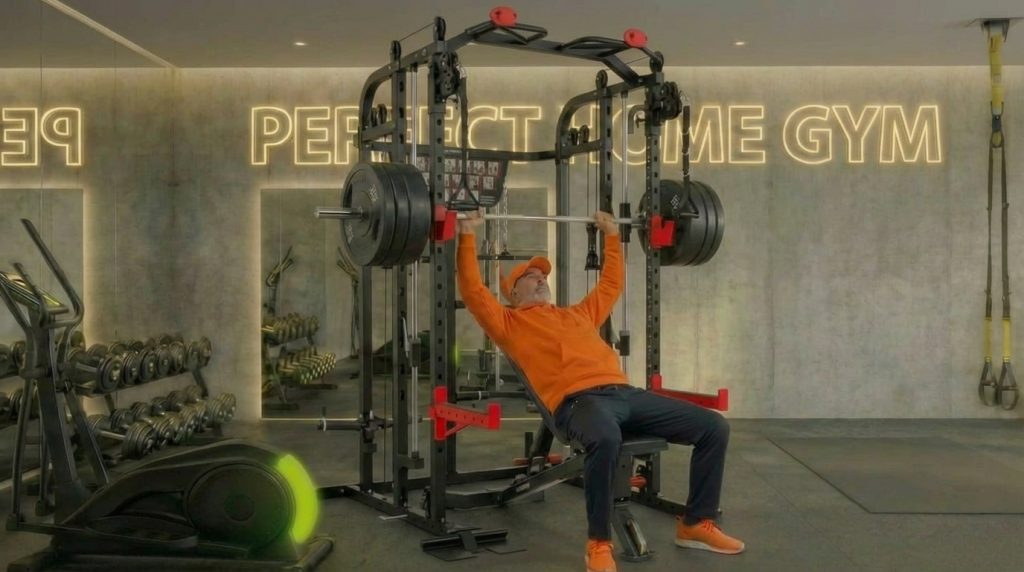 Mann in orange trainiert Bankdrücken in Fitnessstudio mit Perfect Time Gym Schriftzug.