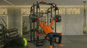 Mann in orange trainiert Bankdrücken in Fitnessstudio mit Perfect Time Gym Schriftzug.