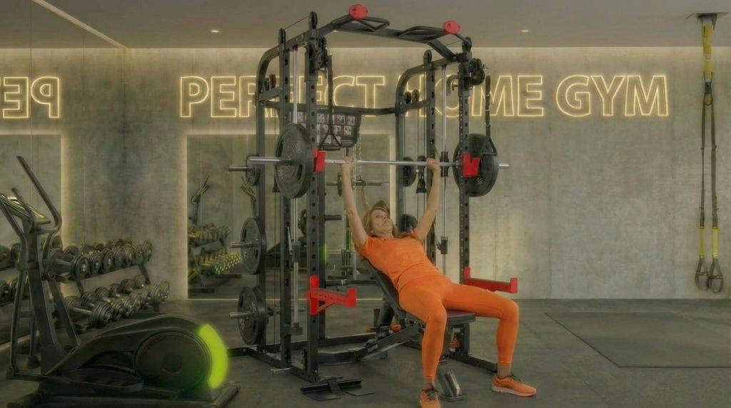 Person in orange Sportbekleidung mit Langhantel auf geneigter Bank im Fitnessstudio mit Geräten und Neonwand.